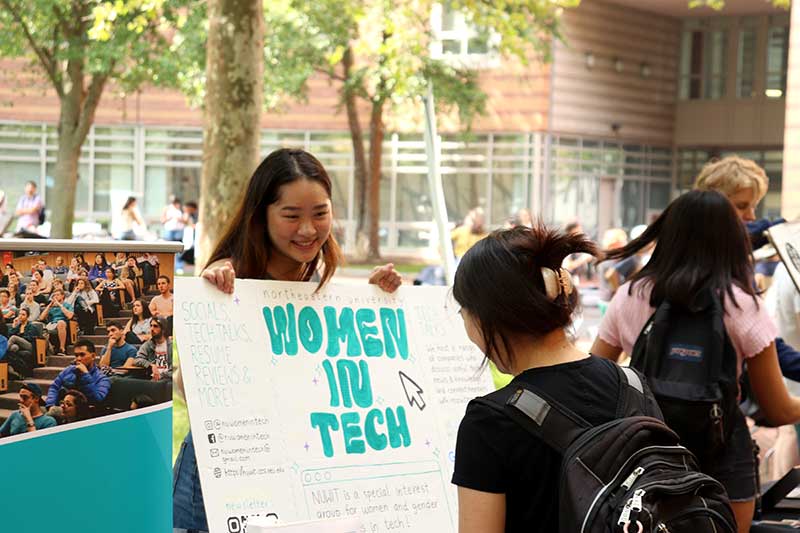 A member of Women in Tech holds the club's poster up while another student asks about the club.