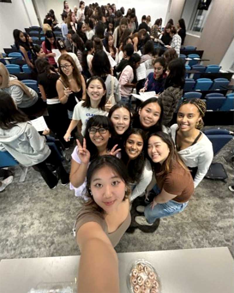 Eight members of NU Women in Technology pose for a selfie an a Northeastern classroom while several other students mill about in the rear of a classroom.