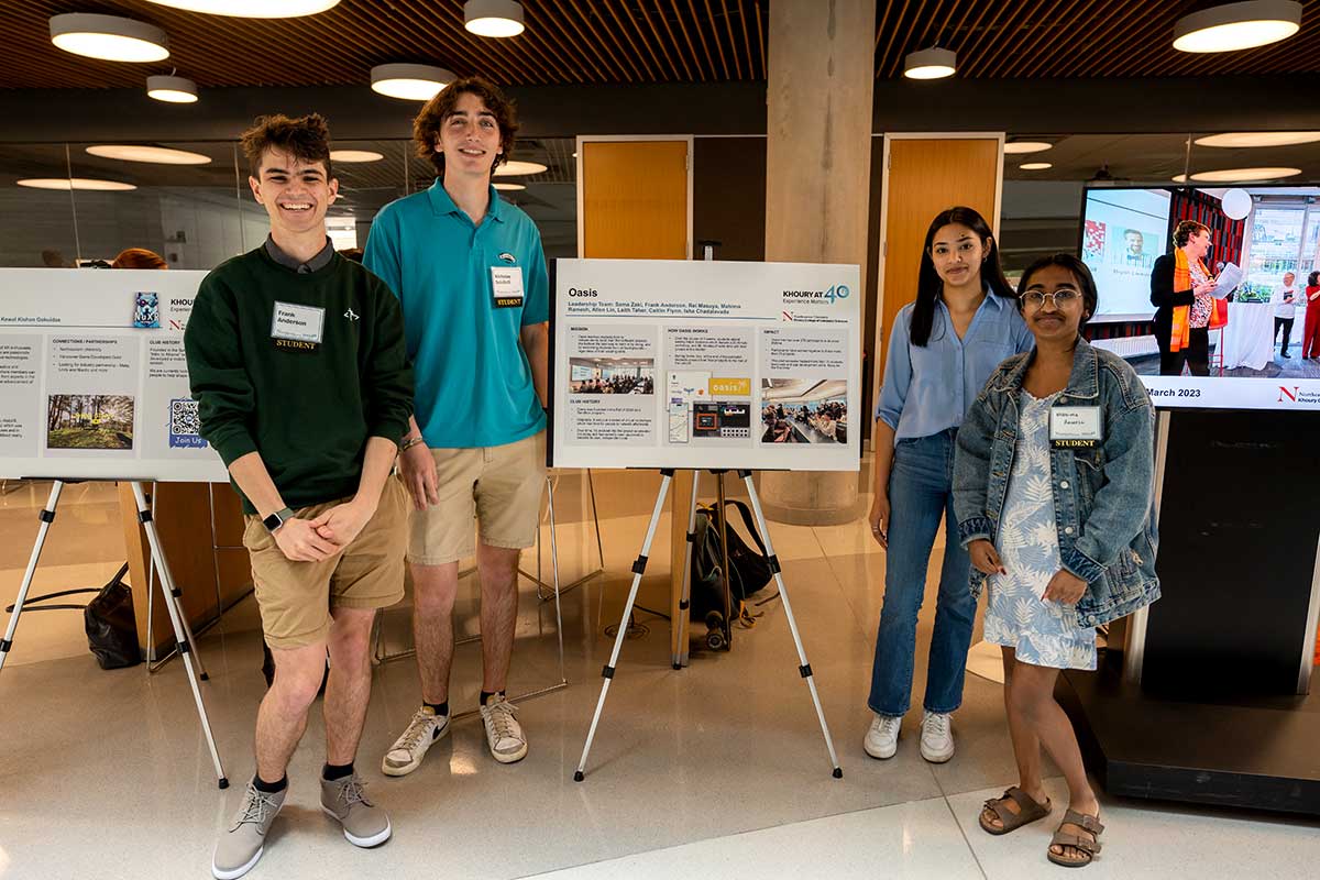 Four members of Oasis stand near their club's poster at a Northeastern club fair in the ISEC building. Two students stand to the left of poster and the other two students stand to the right.