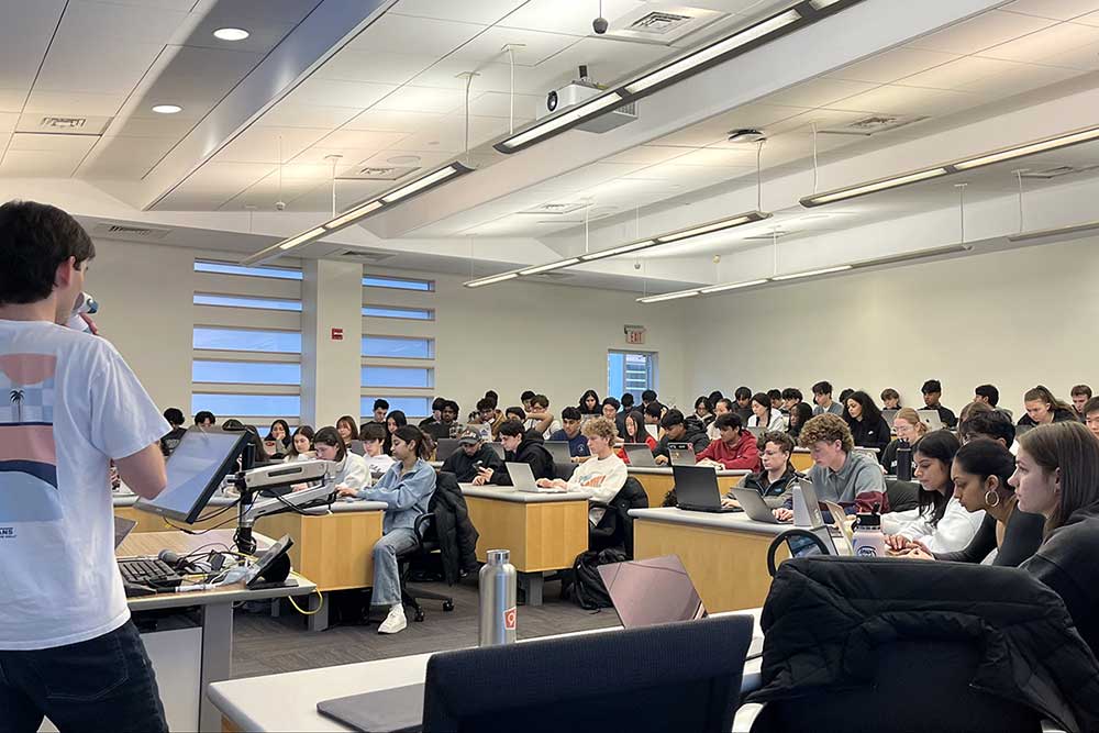 A wide photo of an Oasis meeting in a Northeastern classroom. One student at the front of the classroom is giving a presentation while dozens of other students look on.