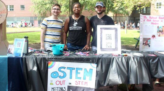 Three members of out in STEM stand behind the club's table at a Northeastern club fair. There are trees and a residence hall in the background.