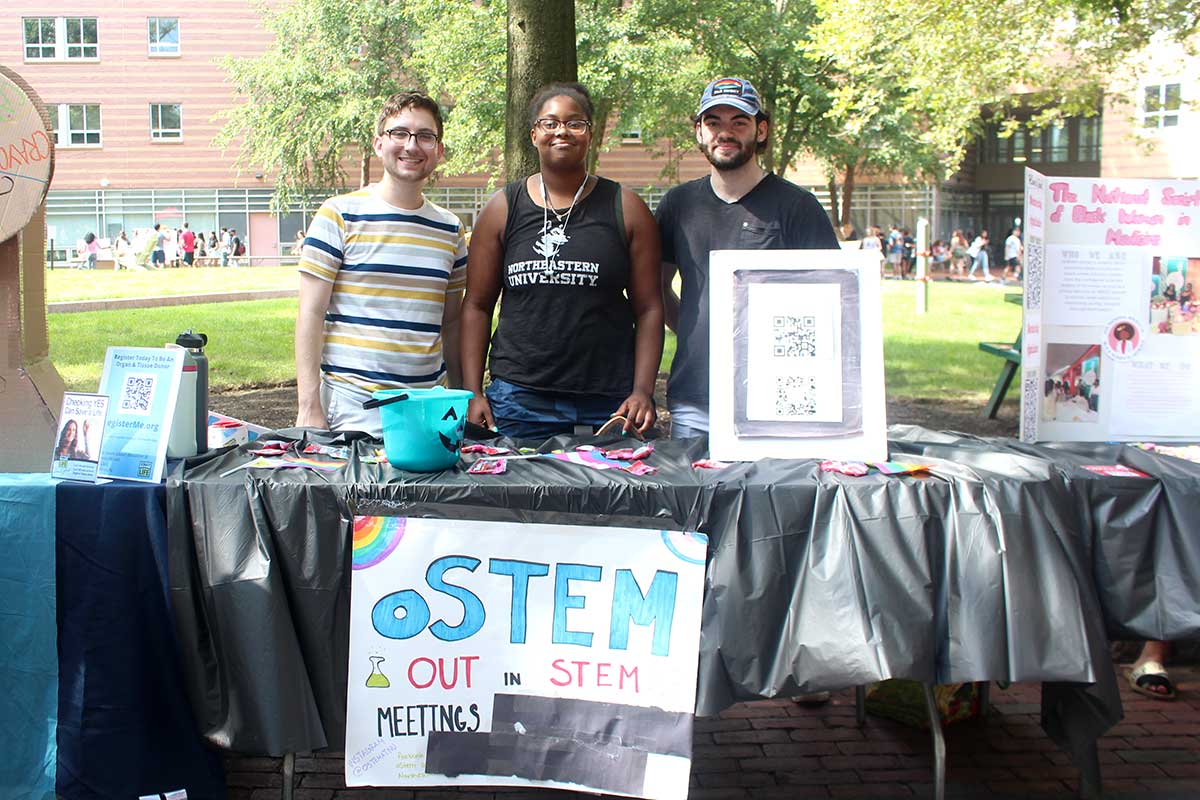 Three members of out in STEM stand behind the club's table at a Northeastern club fair. There are trees and a residence hall in the background.