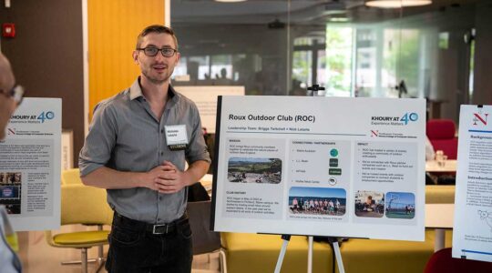 A member of the Roux Outdoor Club stands to the left of his club's poster, which is displayed on an easel.