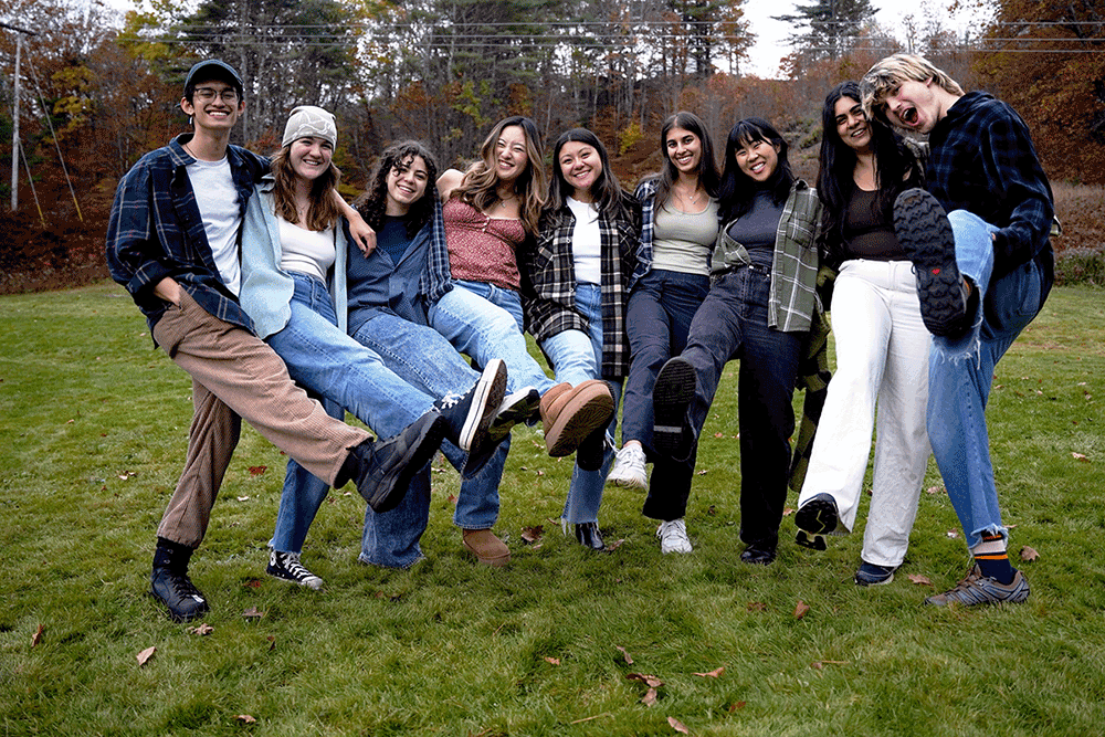 Nine members of Scout pose for a photo while kicking their legs in the air in an outdoor field with trees in the background.