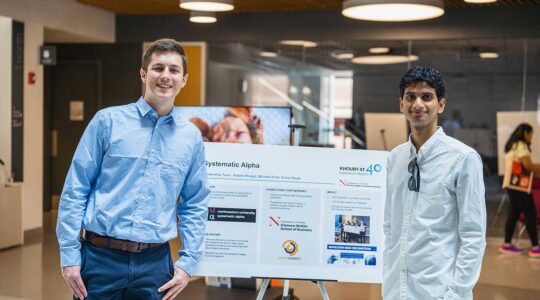 Two members of Systematic Alpha stand on either side of the club's poster inside Northeastern's ISEC building.