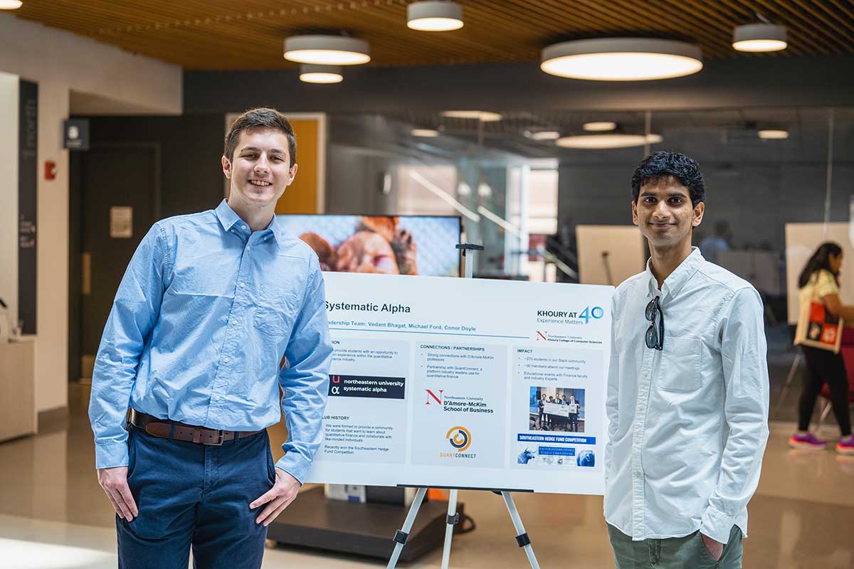 Two members of Systematic Alpha stand on either side of the club's poster inside Northeastern's ISEC building.