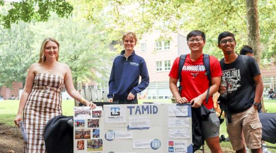 Four members of TAMID pose for a photo standing behind their club's poster. They are standing in a courtyard on the Northeastern campus with trees and a residence hall in the background.