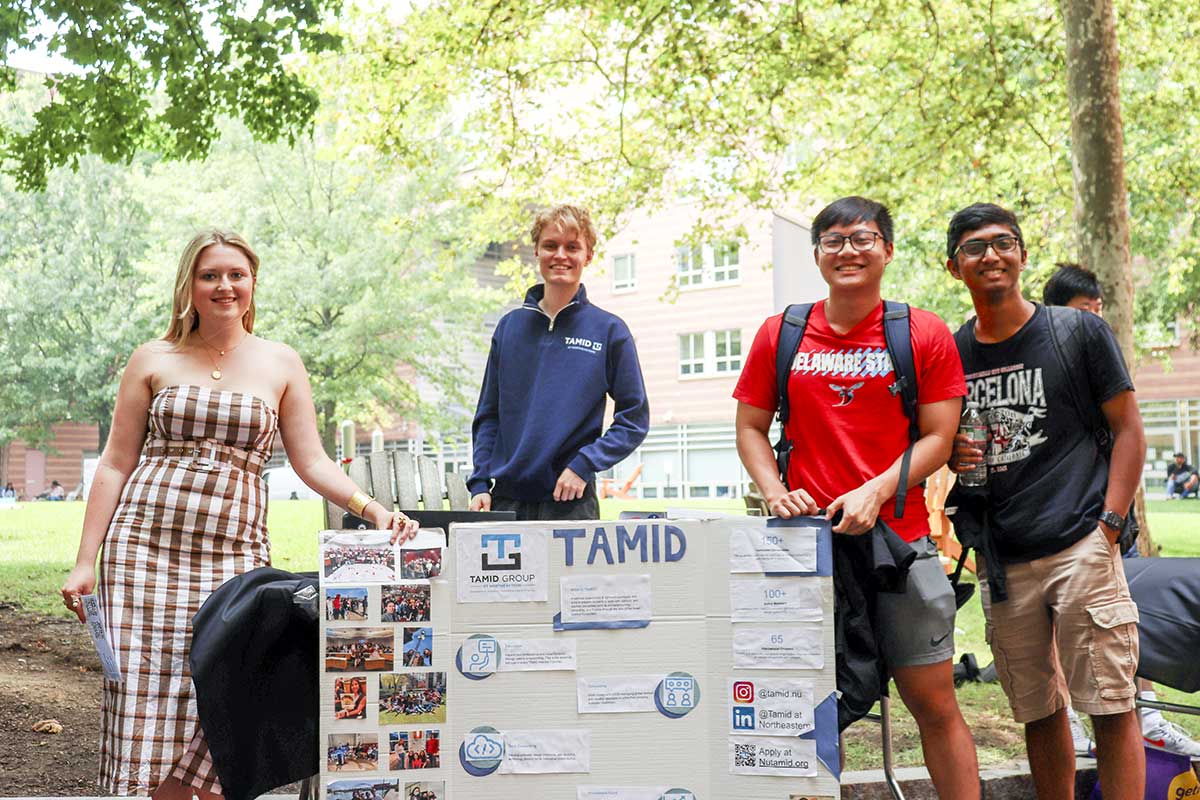 Four members of TAMID pose for a photo standing behind their club's poster. They are standing in a courtyard on the Northeastern campus with trees and a residence hall in the background.