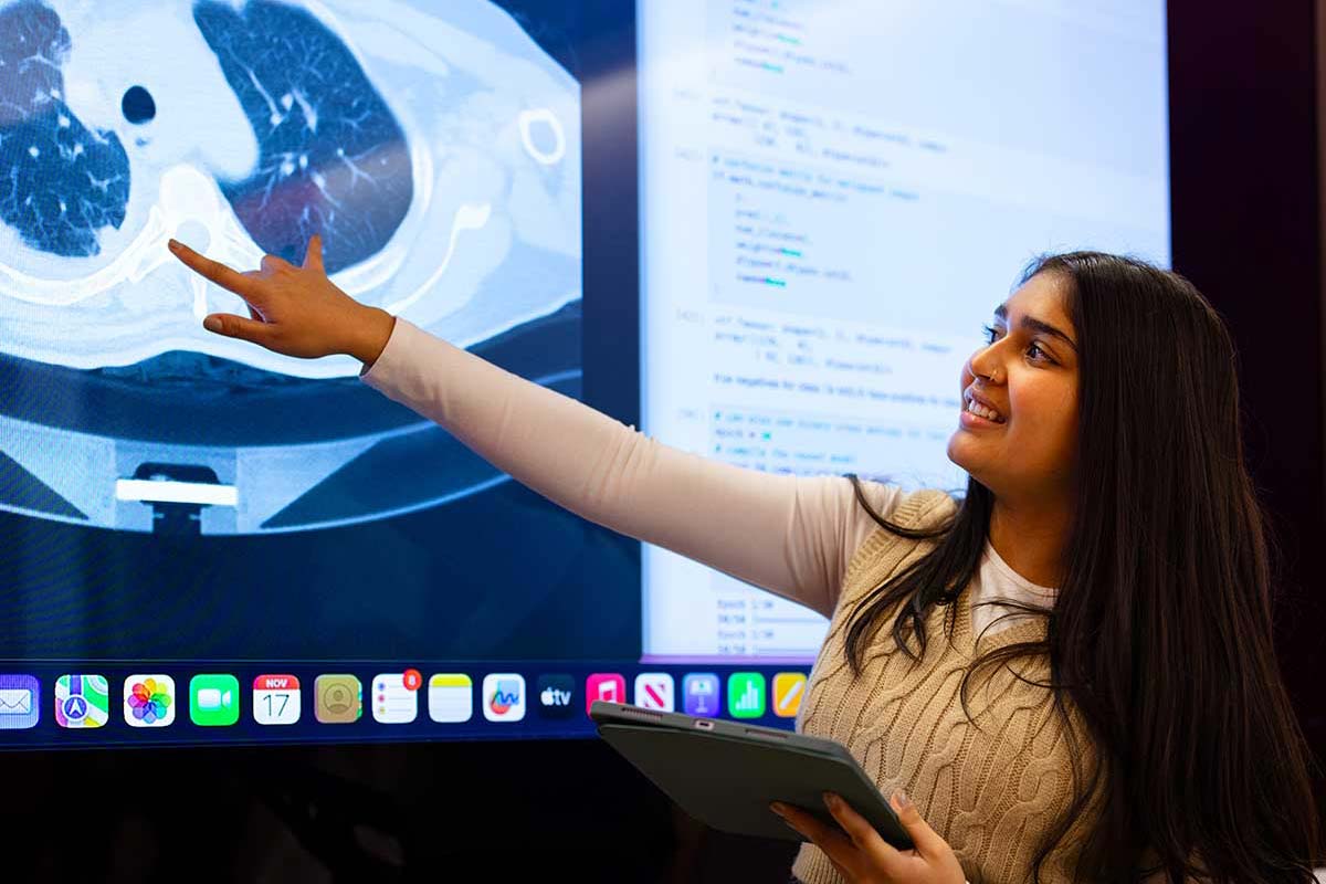 An undergraduate student holding a tablet points to an image projected on a screen. Computer code is visible on the right half of the screen.