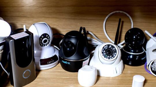 A wooden shelf holds eight networking devices. The networking devices have antennas. Three of the devices are black and the others are white.