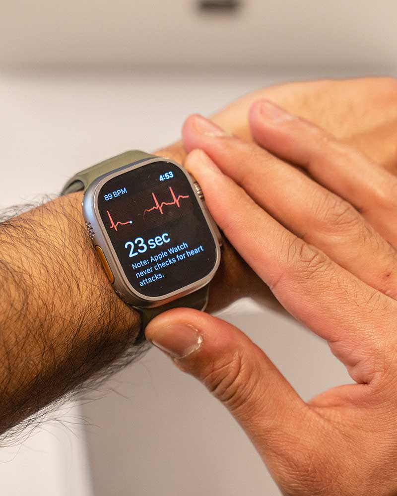 A close-up view of a smart watch on a person's wrist. The person is pressing a button on the right side of the watch face. The watch shows a graph that appears to be monitoring heart rate and the text '23 sec'