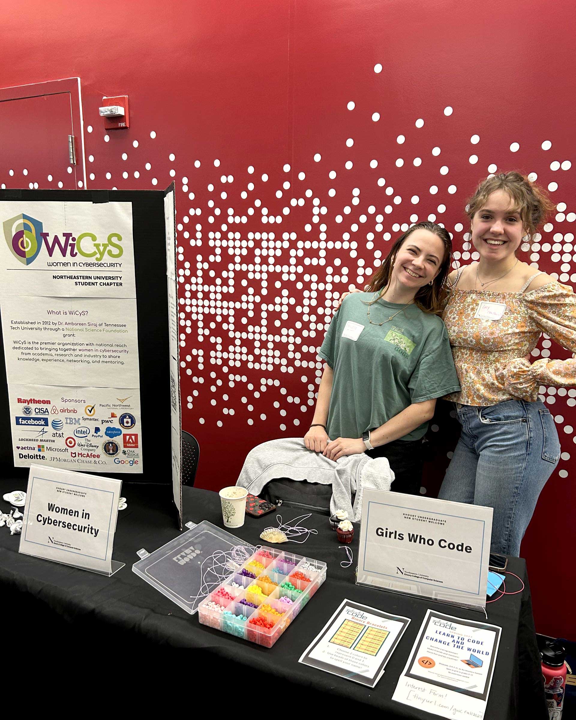 Two members of Women in Cybersecurity pose behind the club's table while smiling. The table has the club's poster, which explains how Women in Cybersecurity started and shows the club's sponsors.