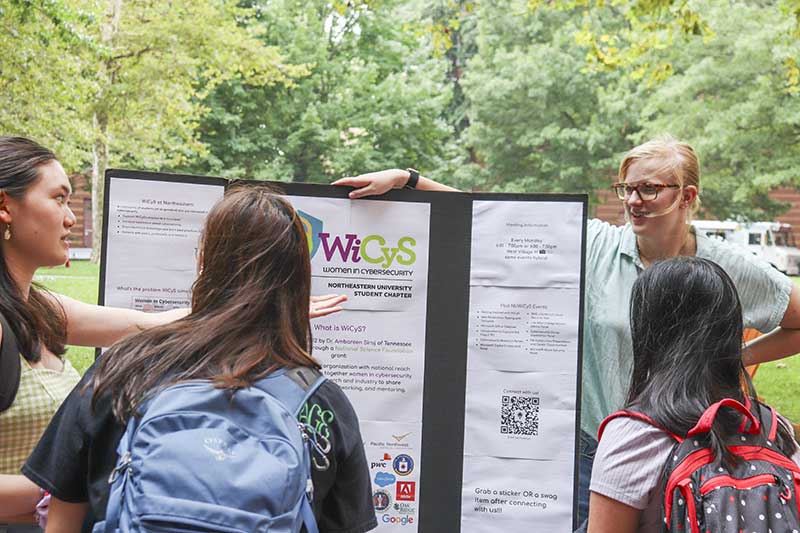 A Women in Cybersecurity members stands next to the club's poster while three students look at the poster. The four students are discussing the club.