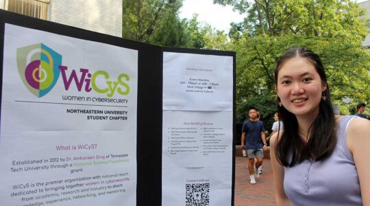 A member of Women in Cybersecurity stands next to the club's poster. The poster shows the logo, explains what the club is, and shows past club events.