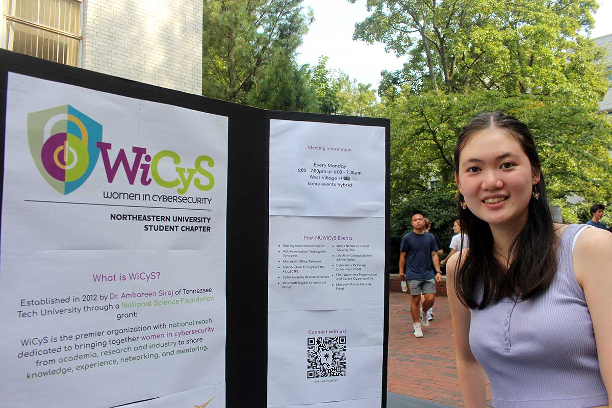 A member of Women in Cybersecurity stands next to the club's poster. The poster shows the logo, explains what the club is, and shows past club events.