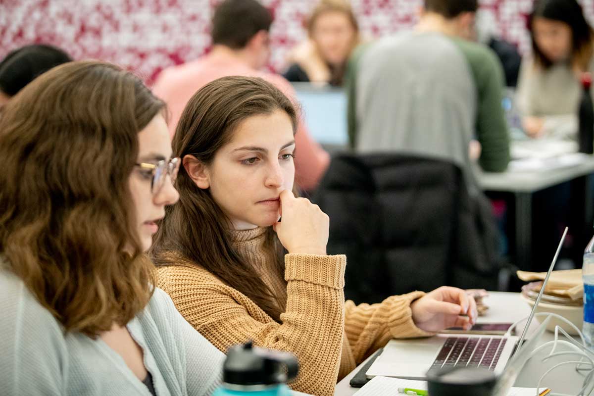 Two women sit at a gray table in Khoury's 102 West Village H lab. The women are looking at the same laptop. The student at left is speaking while the student at right has her right hand under her chin. In the background, four students are seated around a table.