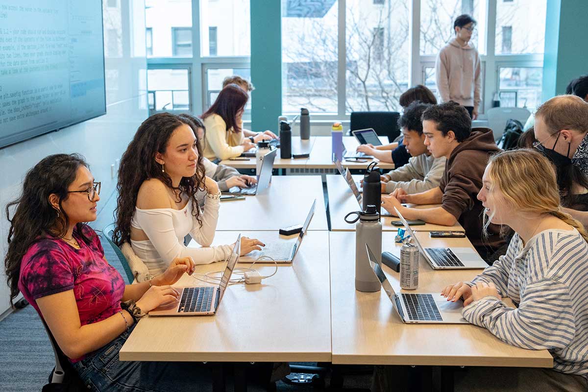 In a Northeastern lab, students sit around four tables that are pushed together. In the foreground, three students talk with their laptops open on the table.