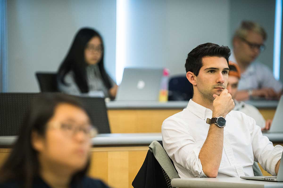 Students watch a lecture in a Northeastern lecture hall. In the right of the photo, one student wearing a while shire sits with is elbow on the table and his hand under his chin.