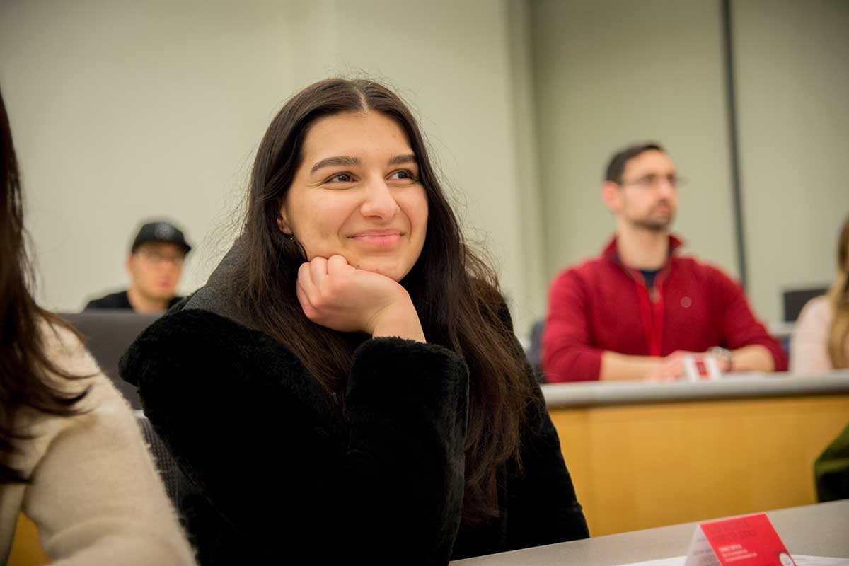 A student wearing a black jacket seated in a Northeastern lecture hall smiles while watching a presentation. There are two students seated in the room's back row in the background.