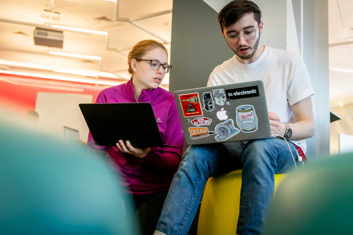 Two students collaborate on a project in the 102 West Village H Lab. The student on the left is standing while leaning agains a pillar and holding an open laptop in her left hand. The other student is seated on a soft stool while looking at his laptop, which is open on his lap.