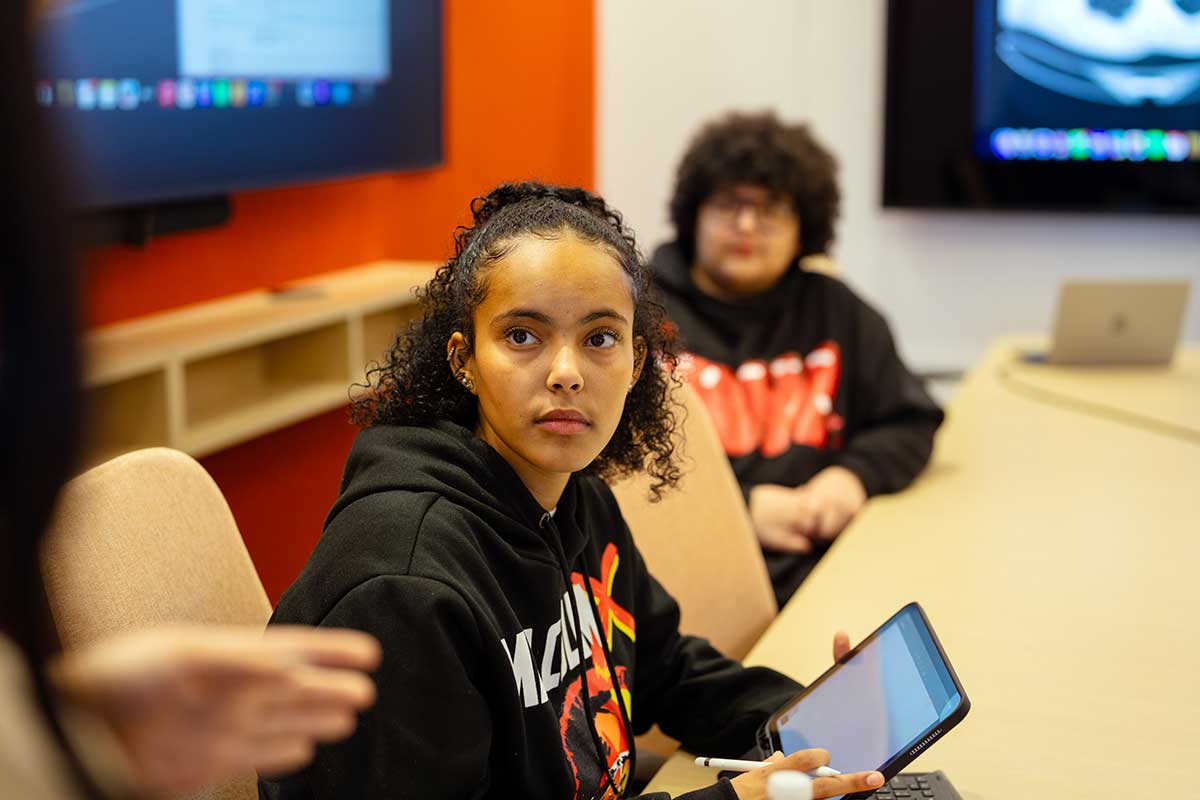 In a Northeastern classroom, a student sits at a table and listens attentively. There are large screens on the two walls in the background.