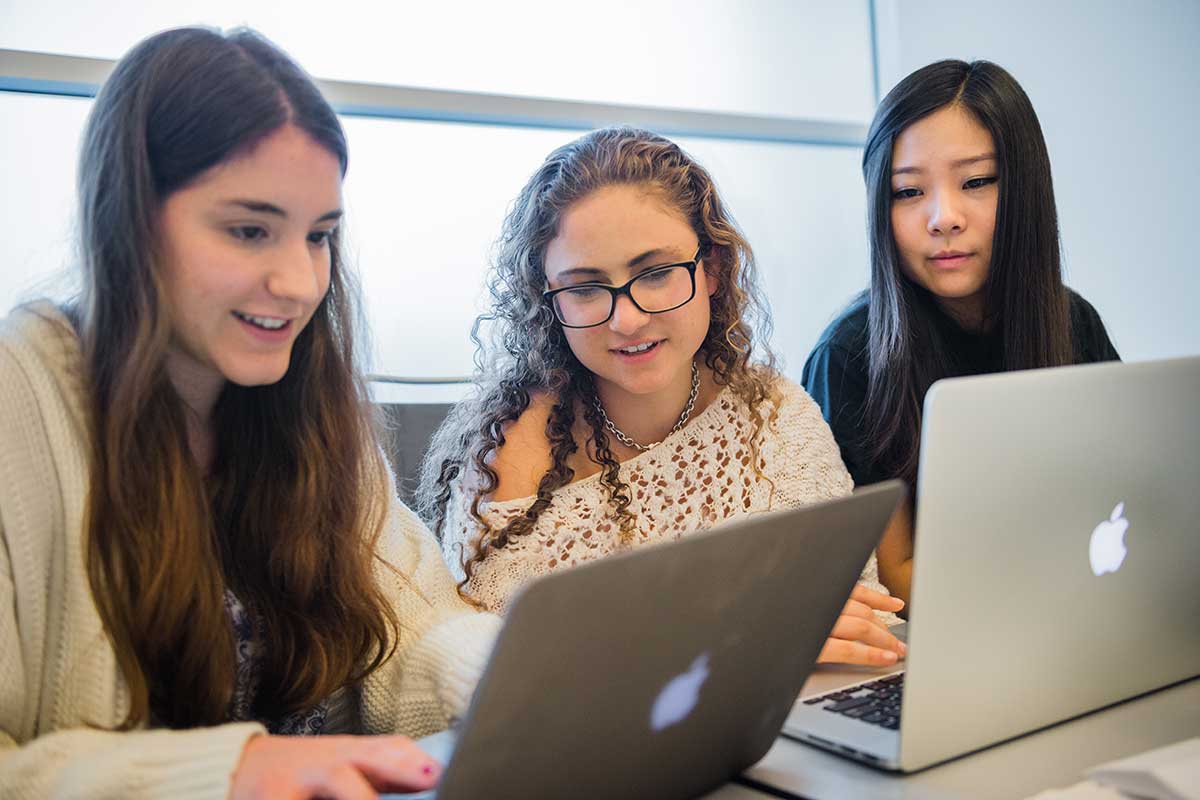 In a classroom, three students look at a laptop and discuss an assignment.