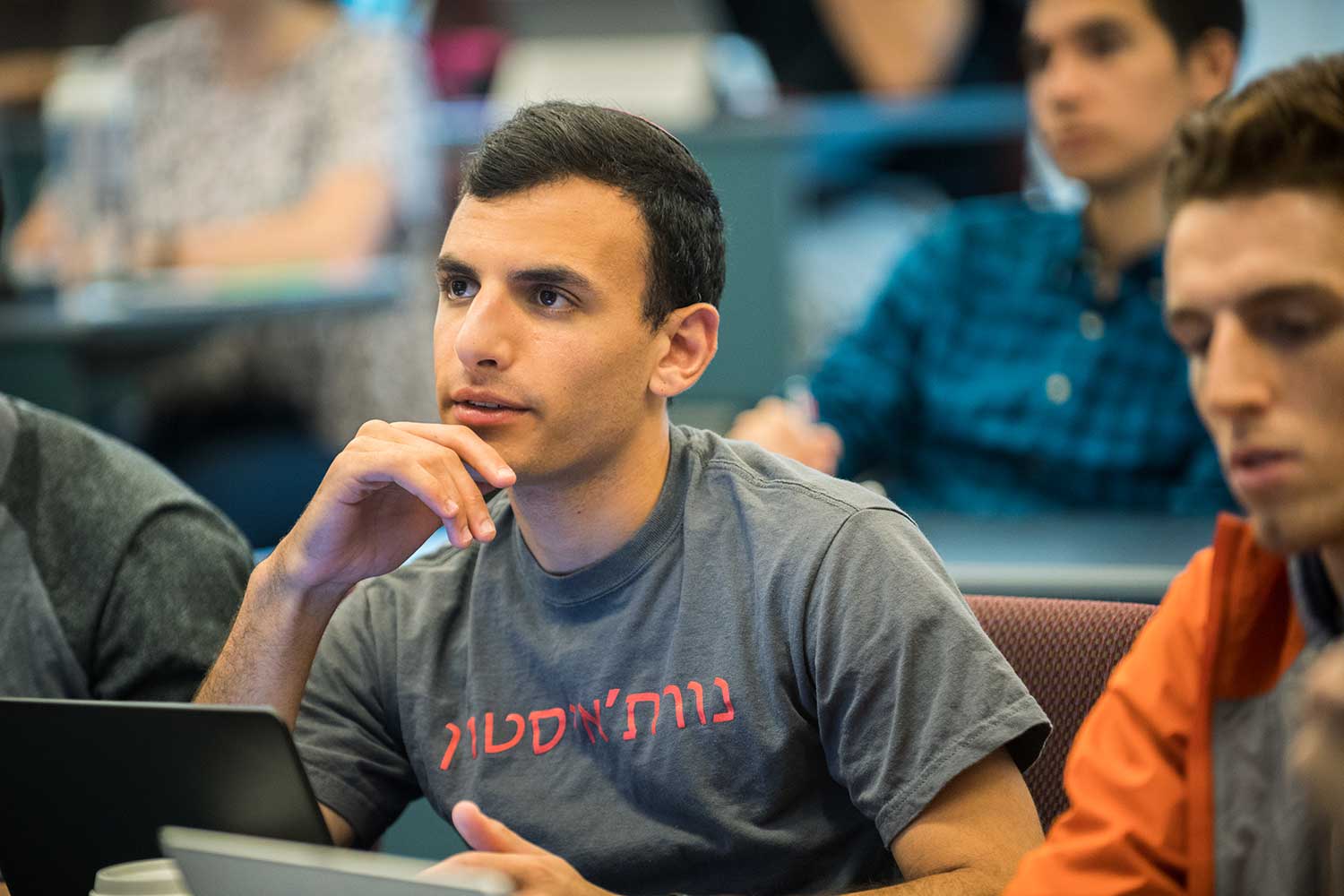 A student watches a lecture attentively in a Khoury College classroom. A laptop is open on the table in front of him. To the right, another student looks down at the table.