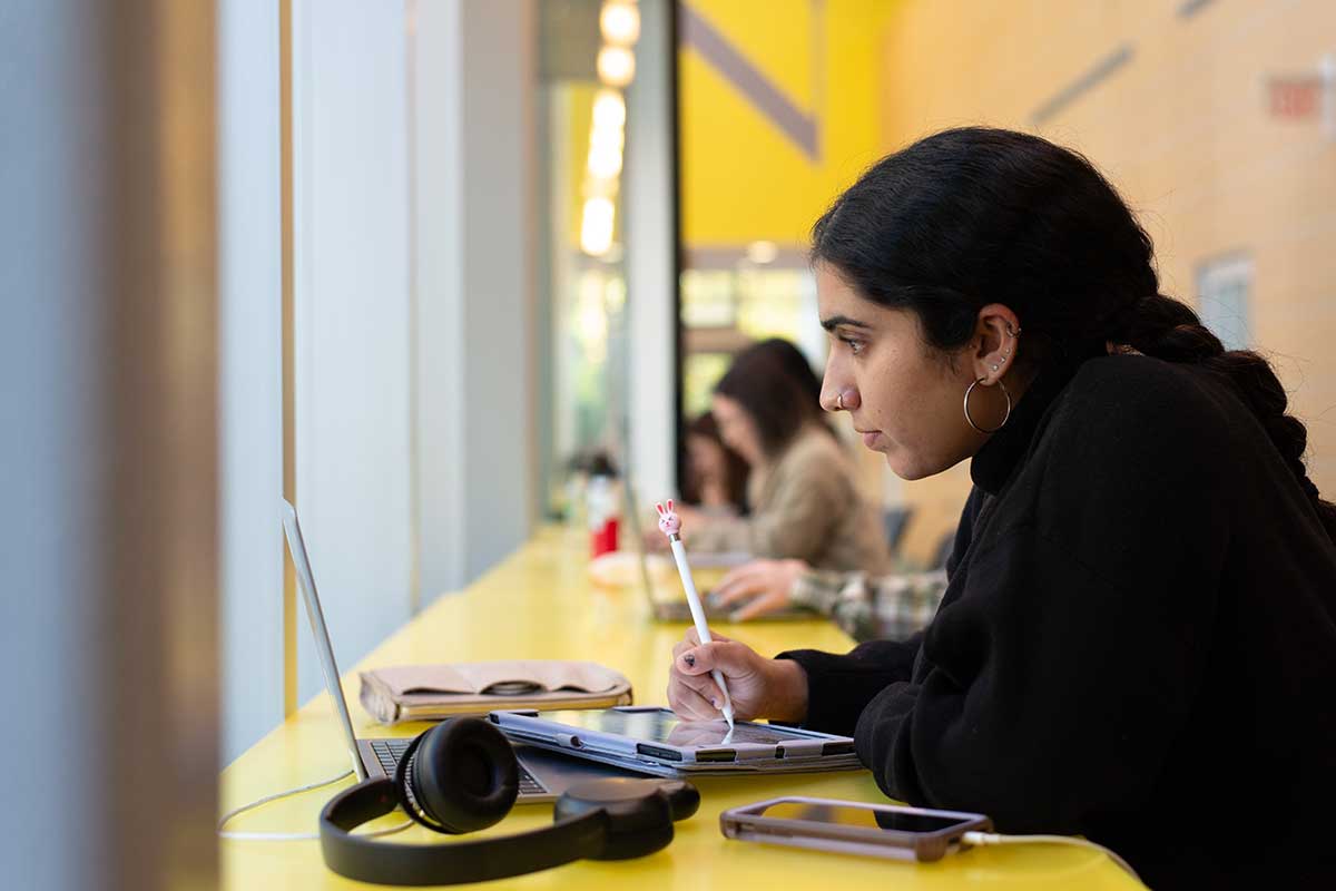 In the atrium of West Village H, a student writes on a tablet with a stylus while viewing a laptop screen.