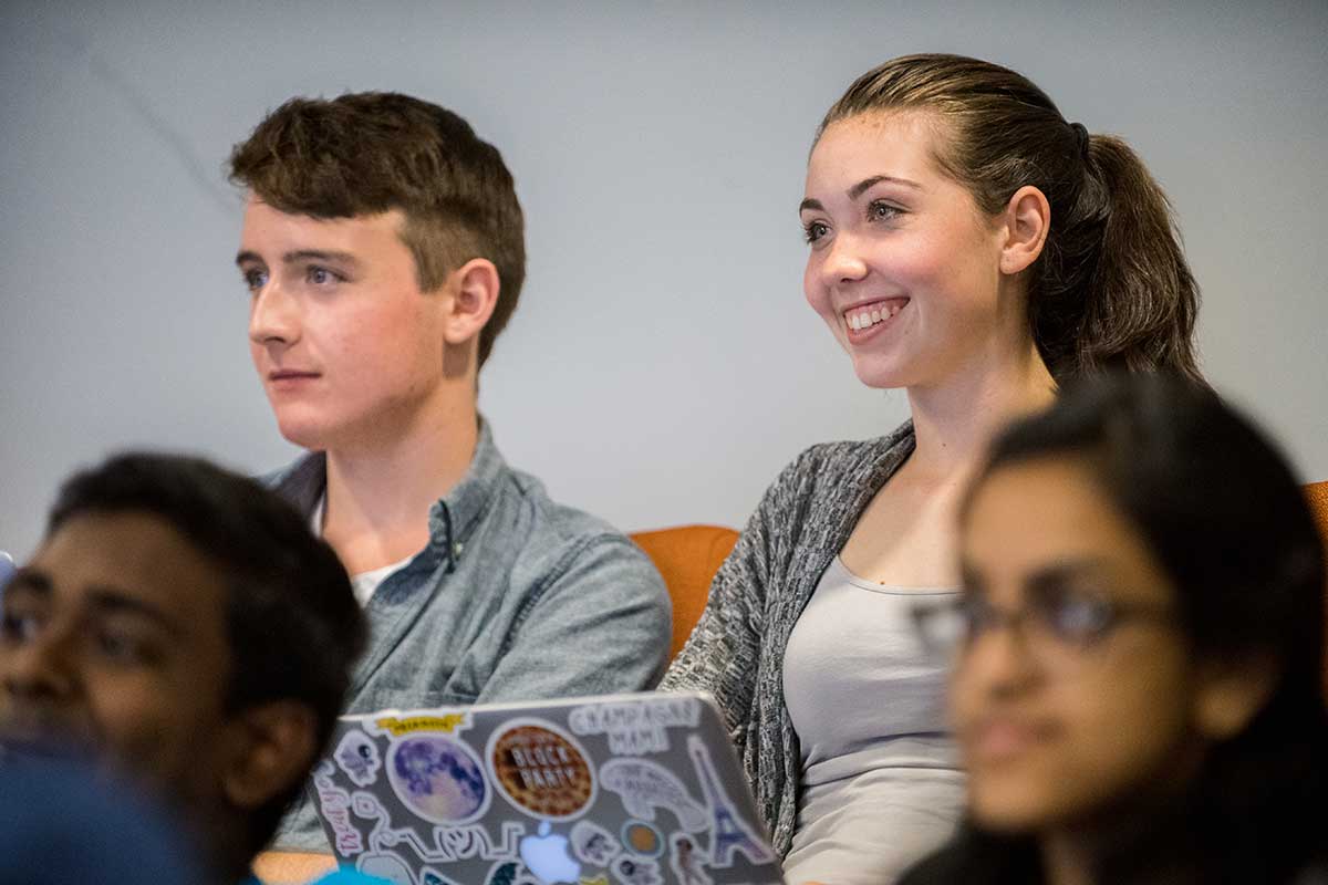 Two students watch a lecture during a course at Khoury College. The students are seated in orange chairs. The student on the right is smiling widely.