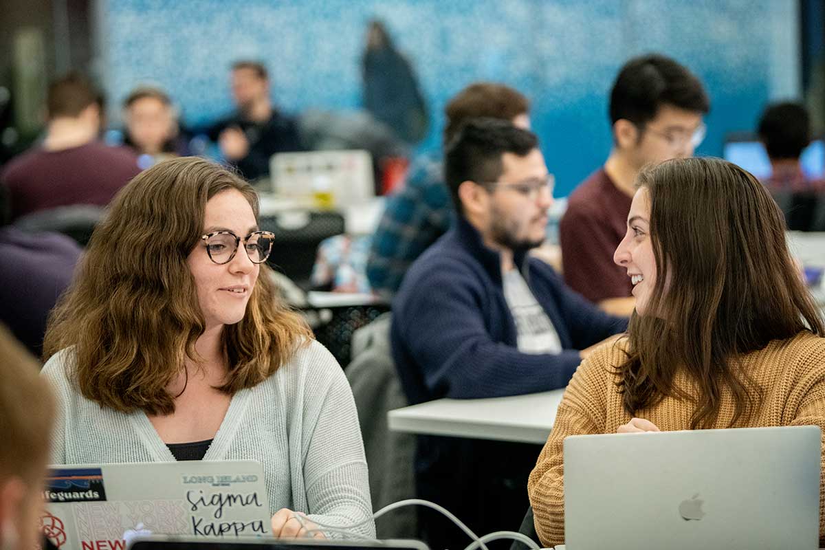 Students work together in a the West Village H computer lab at Northeastern. In the foreground, two students smile during a conversation while their laptops are open on the table in front of them. Other students are sitting at tables in the background.