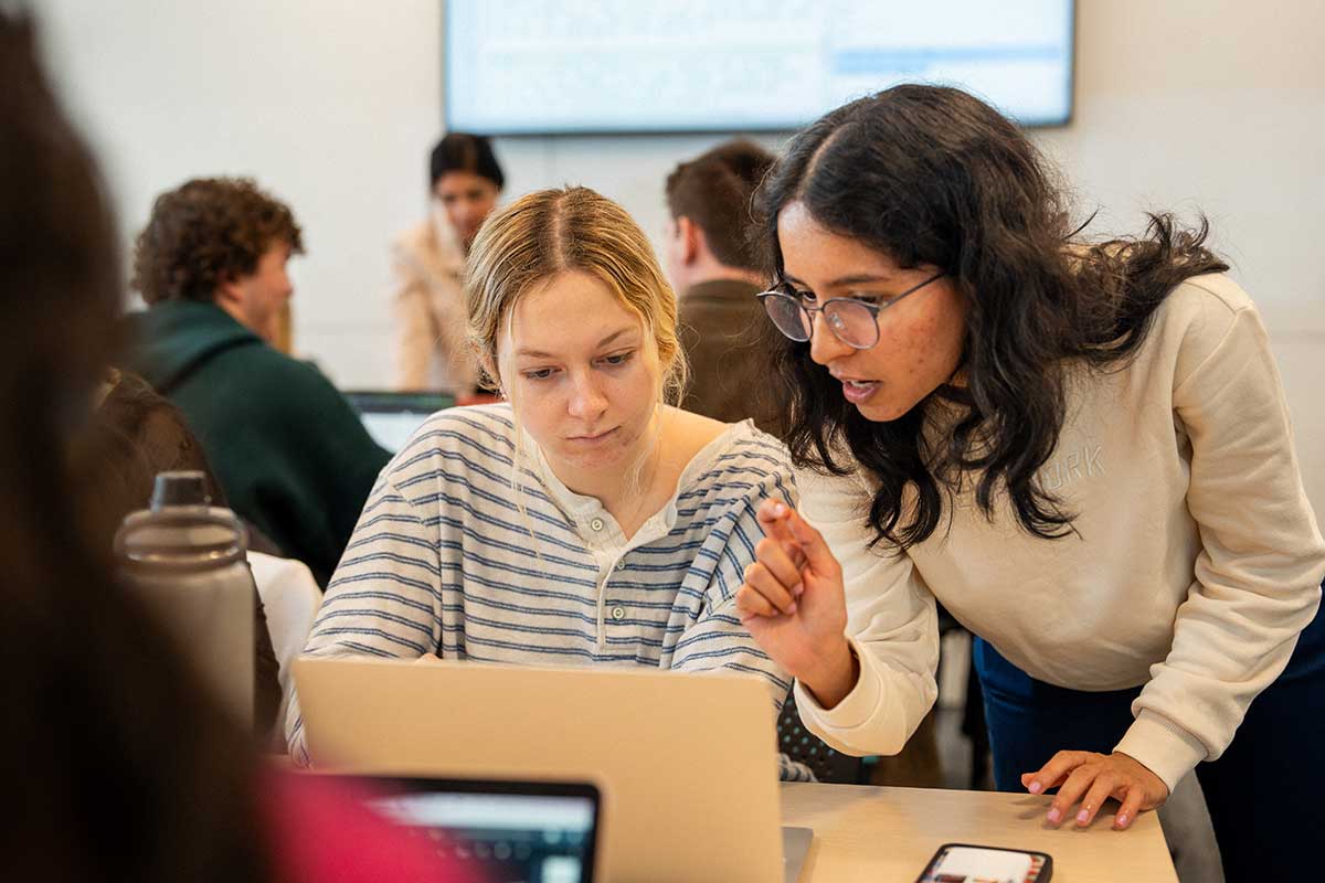 In a Northeastern lab, students sit at tables and work on a project. In the foreground, a student sitting at a table looks at her laptop while a faculty member leans over and explains the exercise.