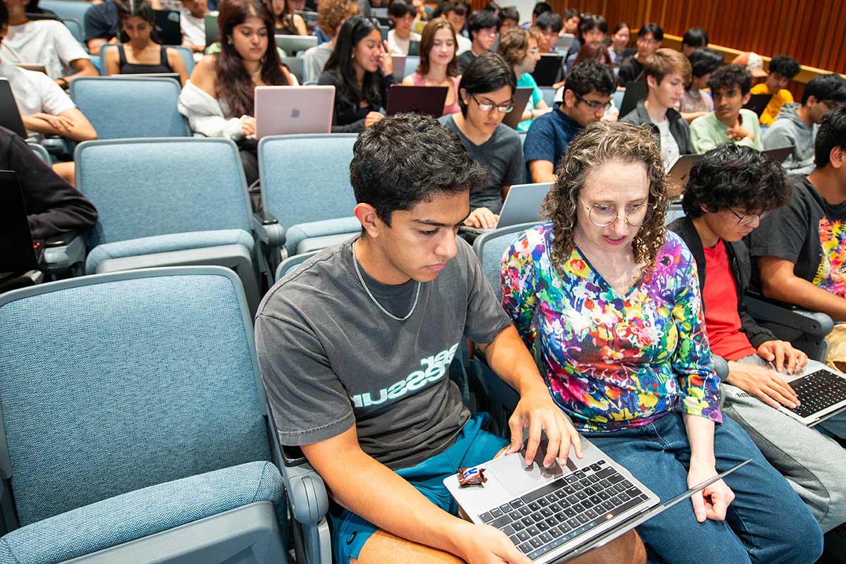 Dozens of students sit in blue chair in a Northeastern lecture hall. In the first row of chairs, a student looks at his laptop and moves his fingers on the trackpad while a Northeastern faculty member explains the assignment.