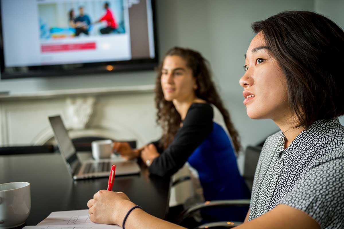 Two students sit in a classroom while an image is projected on a large screen on the wall in the background. The student in the foreground is holding a red pen, and the student in the background has a laptop open on the table.