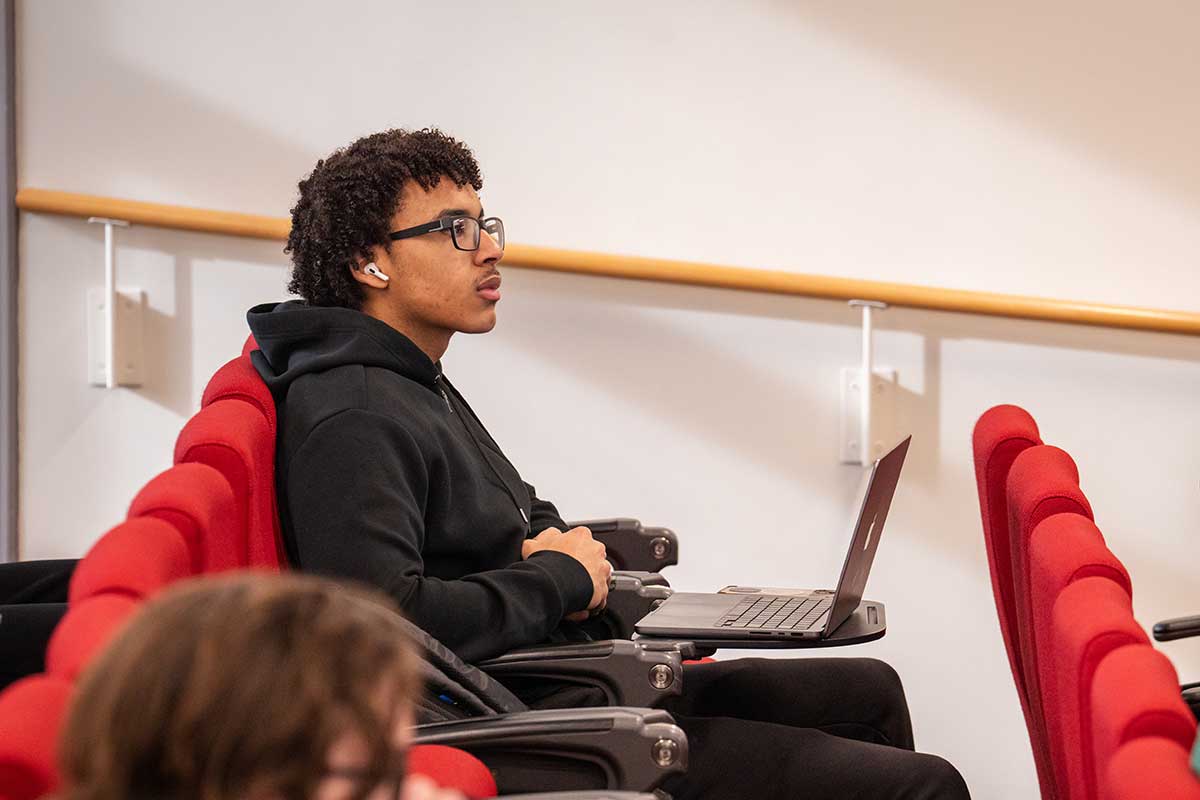 A student sits in a red chair while viewing a class lecture in a Northeastern lecture hall. The student has a laptop open on the desk in front of hime.