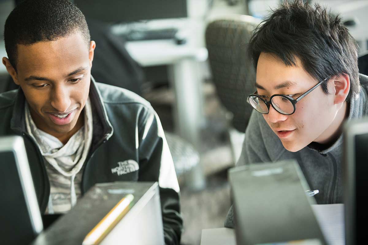 Two students work on a project together in a Northeastern computer lab. The photo is taken from behind a monitor screen and both students are looking at the screen.