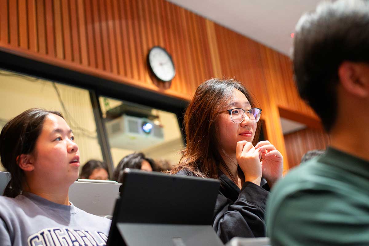 Students view a lecture in a Northeastern lecture hall. the student in the center of the photo has an attentive expression. The walls of the room in the background have wooden paneling.