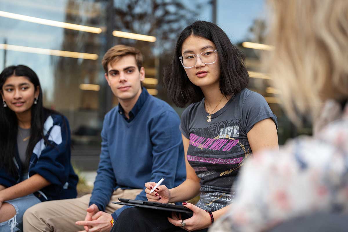 Four students converse in an outdoor courtyard at Northeastern. In the foreground, the back of a student's head is visible facing the other three students in the photo.