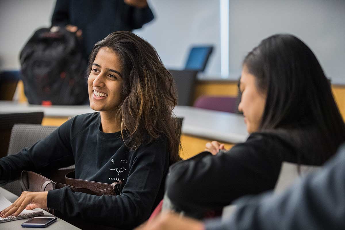 Two students are seated in chairs in a Northeastern lecture hall. The student on the left smiles while viewing a presentation.