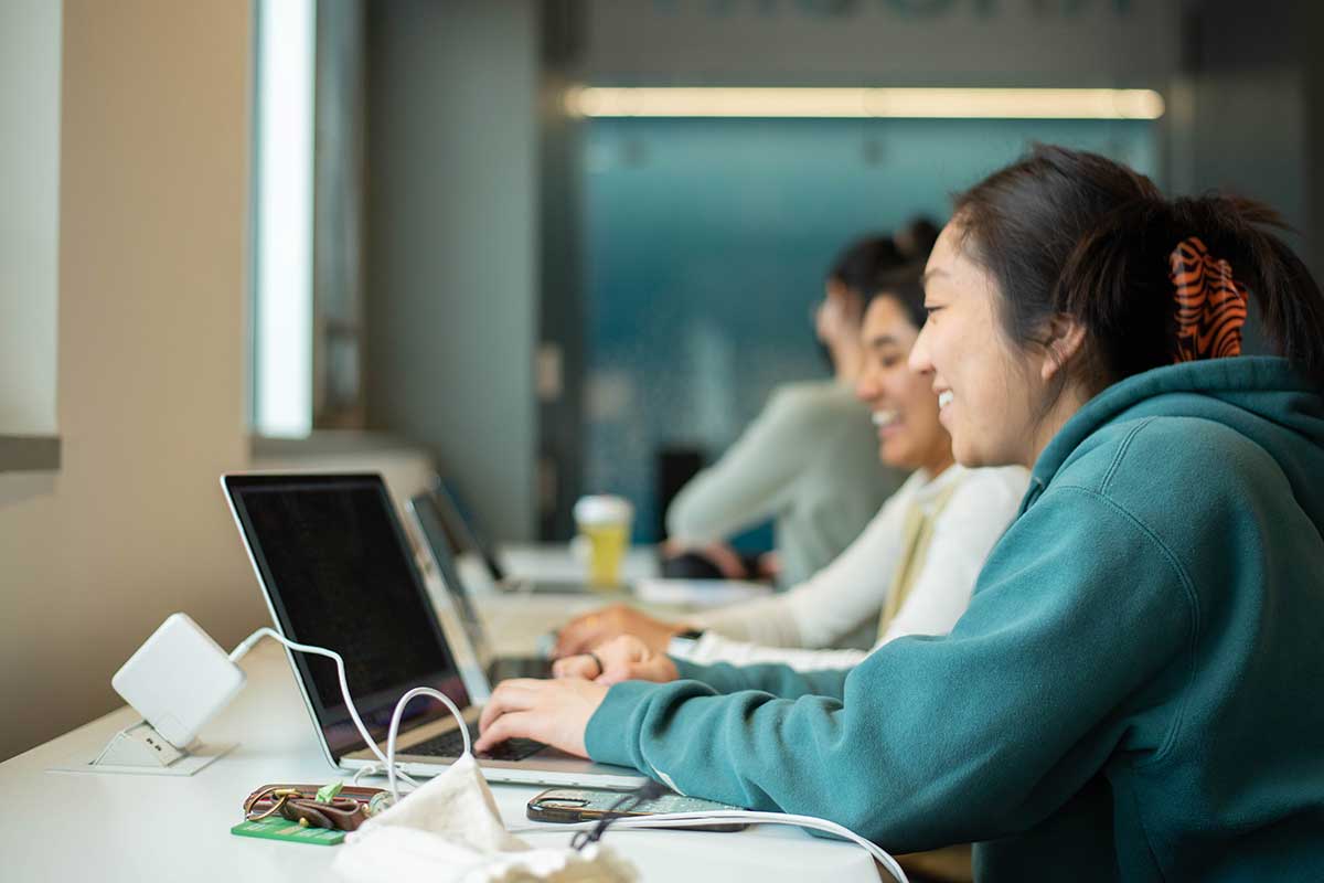 Two students sit at a table in a study area in Northeastern's West Village H. The students are smiling while they view their laptop screens.