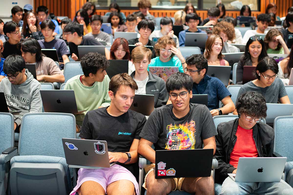 Dozens of students prepare for a course session in a Northeastern lecture hall. The students are separated into about 10 rows of blue chairs.