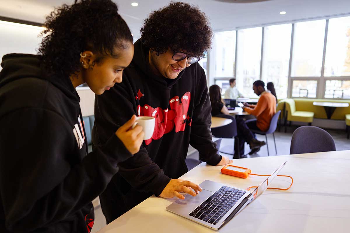 Two students stand in front an open laptop sitting on a table. The student on the right is moving his fingers on the laptop's trackpad, while the student on the left views the laptop screen while holding a mug.