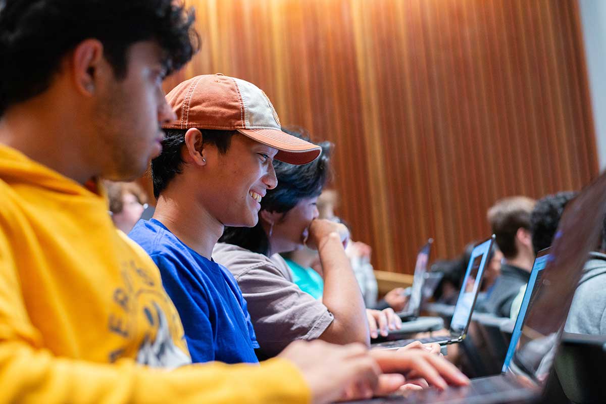 Several students sit in a row of seats in a Northeastern lecture hall. The student in the foreground is smiling as he views his laptop screen.