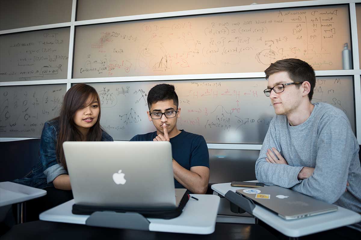 In a West Village H study area, three students sit at mobile desks while looking at a laptop open in front of the student sitting in the middle. In the background, a wall is covered with written mathematical equations.