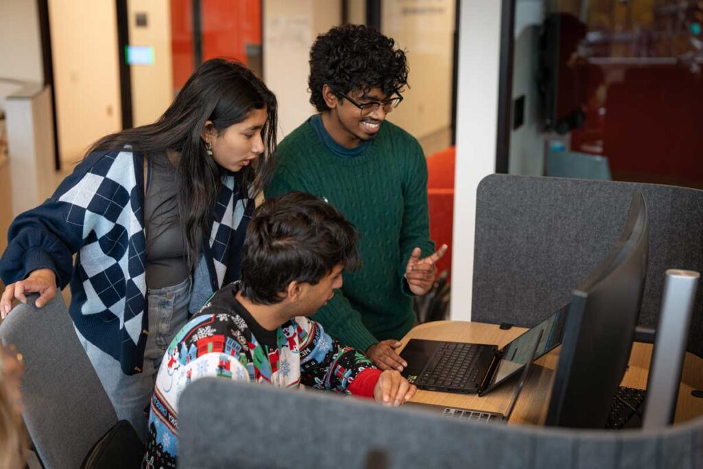 Three students look at a laptop screen sitting on a desk. Two students are standing and one student is seated.