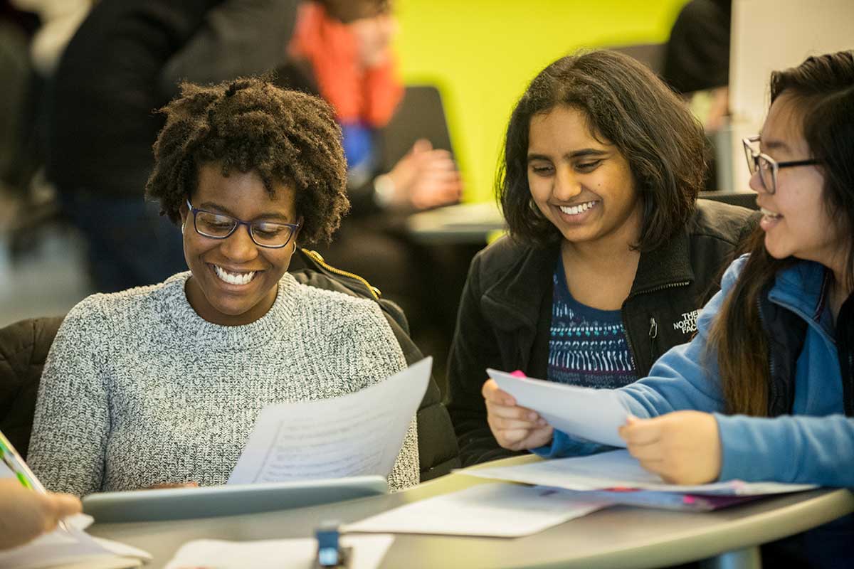 Three students smile and laugh while they sit around a small table looking at a piece of paper.