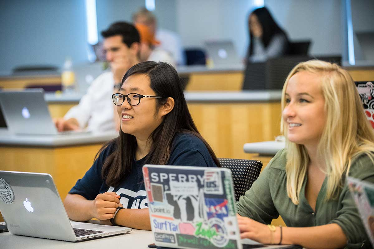 Students watch a lecture in a Northeastern classroom. In the foreground, two students listen attentively with their laptops open on the table in front of them.