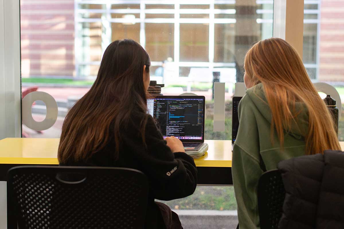 A photo taken behind two students who are seated at a table in the West Village H atrium looking at a laptop. The students are facing windows through which a campus courtyard is visible.