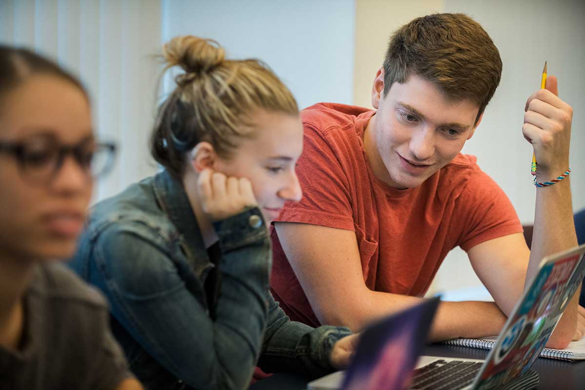 Two students view a laptop while seated in a Northeastern lecture room. To the right of the photo, another student views her laptop.