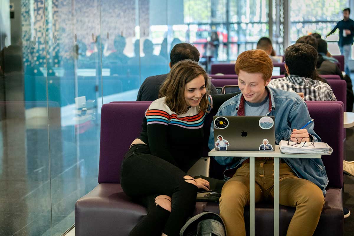 Two smiling students seated on a purple bench view a laptop computer while working on a project in the West Village H 102 lab.