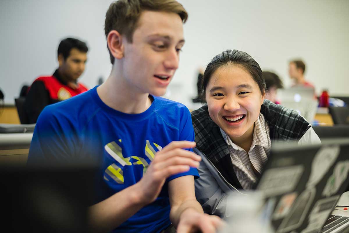 Two students work on a project during a class session. The students are looking at an open laptop. One student is speaking and the other is smiling widely.